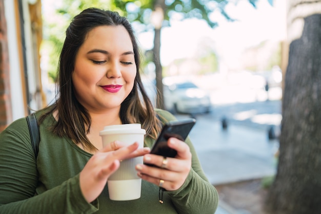 Portrait of young plus size woman typing text message on her mobile phone while holding a cup of coffee outdoors at the street