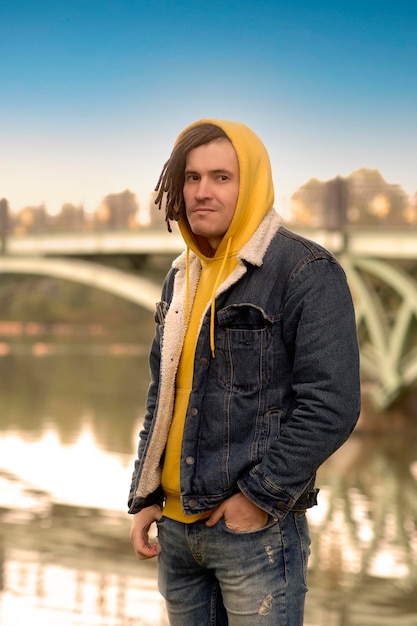 Portrait of young positive man with dreadlocks in hood posing in city park on background of blurred bridge in cloudy weather