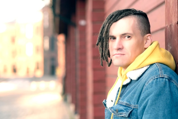 Portrait of young positive man with dreadlocks standing leaning on wall on city street