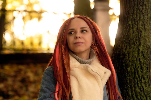 Portrait of young positive woman with ginger dreadlocks looking around standing near tree trunk