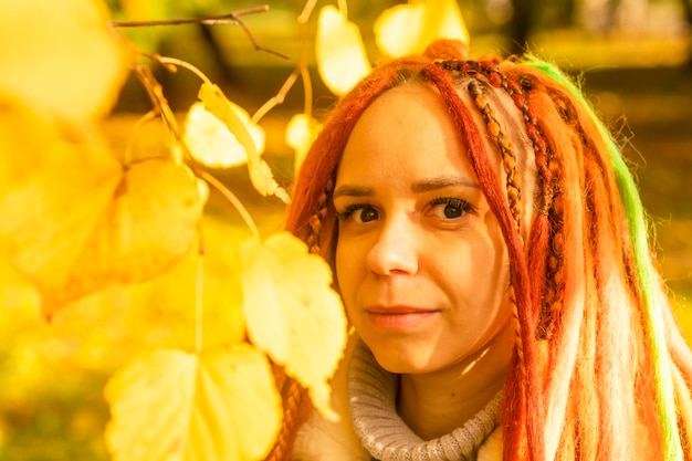 Portrait of young positive woman with ginger dreadlocks standing among branches of tree with yellow leaves