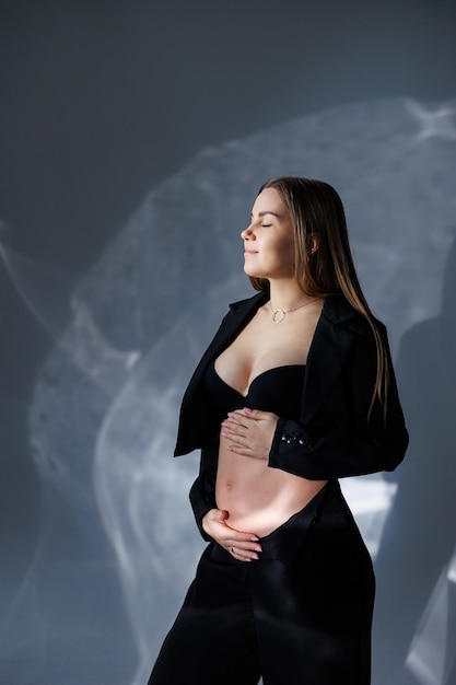 Portrait of a young pregnant woman in a black classic suit A pregnant woman on a gray background hugs her belly