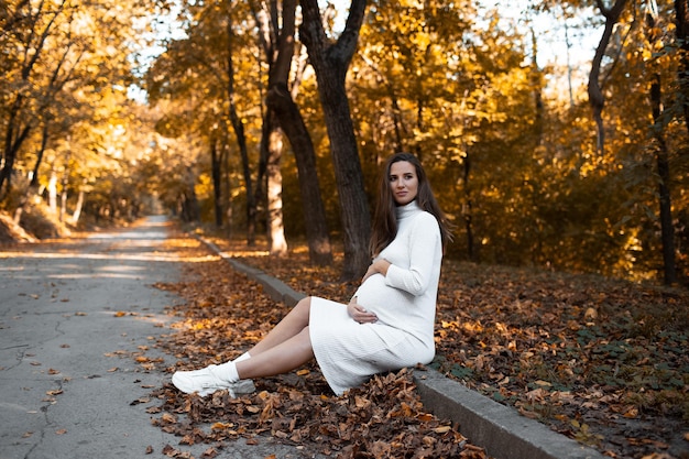 Portrait of young pregnant woman touching the belly with hands sitting on the border in park wearing white dress on background of golden autumn trees