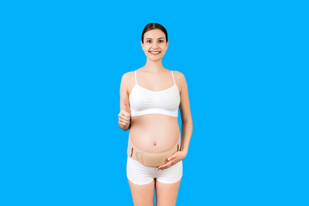 Portrait of young pregnant woman in underwear wearing bandage on her belly and showing thumb up sign at blue surface with copy space