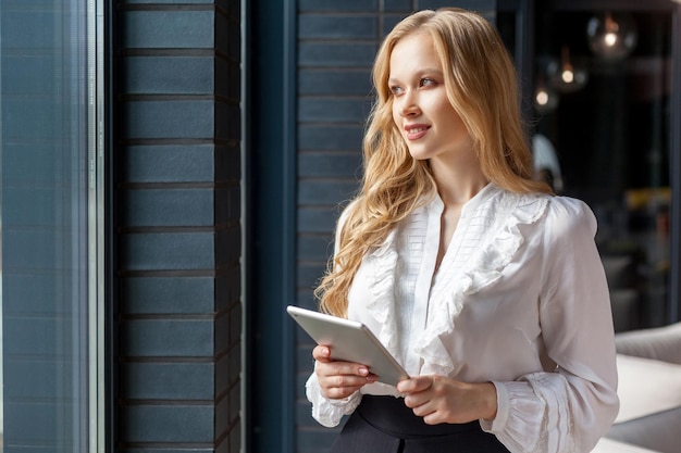 Photo portrait of young pretty businesswoman with blond hair in stylish classic shirt looking out window and smiling holding tablet pc digital device for office work business applications indoors