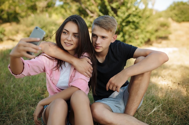 Portrait of young pretty couple sitting on lawn in park and making selfie together. Cute couple taking photos on cellphone frontal camera