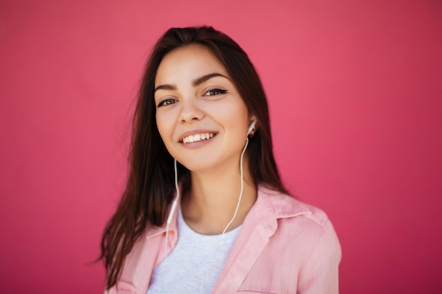 Portrait of young pretty smiling girl with dark hair standing and listening music in earphones while happily looking in camera on pink background