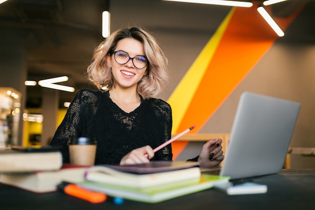 Portrait of young pretty woman sitting at table in black shirt working on laptop in co-working office, wearing glasses