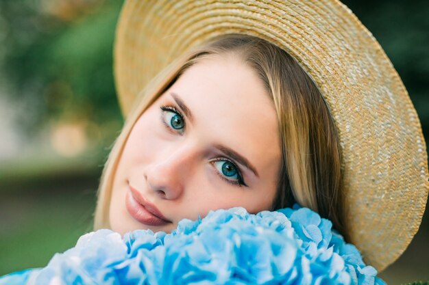 Portrait of young pretty woman in straw hat with face near hydragea flower on the greeen street. Pure beauty.