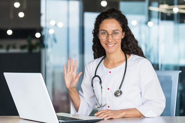 Portrait of a young professional Latin American female doctor in a white medical coat smiling and looking at the camera holding her hand up gesture of greeting the female doctor is working inside