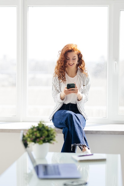 Portrait of young redhaired smiling woman sitting on windowsill with phone