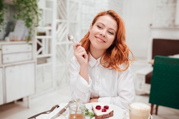 Photo portrait of young redhead pretty woman eating cake in cafe while drinking coffee.