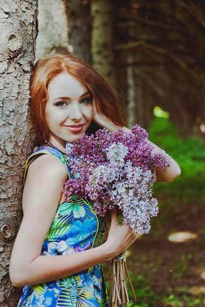 Portrait of young redhead woman in spring garden. freckles. blooming purple flowers. Lilacs bouquet