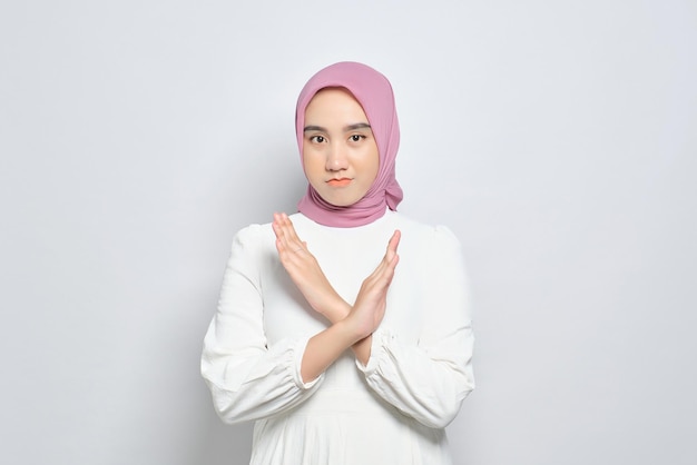 Portrait of young serious Asian Muslim woman showing stop gesture with crossed arms isolated over white background