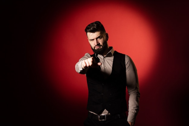 Portrait of a young serious bearded man in a black vest on red background. Man pointing at the camera