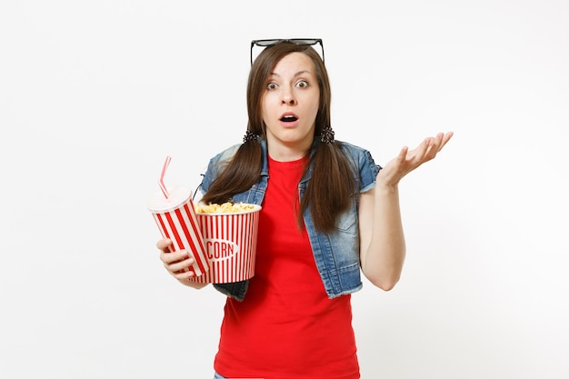 Portrait of young shocked woman in 3d glasses watching movie film, holding bucket of popcorn, plastic cup of soda or cola spreading hands isolated on white background. Emotions in cinema concept.