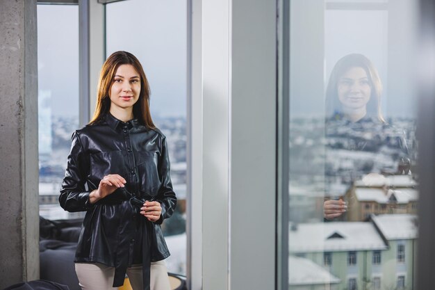 Portrait of a young slender woman in glasses and a black leather shirt Modern woman on the background of the window in the office with a large window