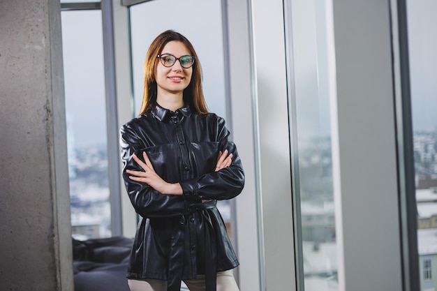 Portrait of a young slender woman in glasses and a black leather shirt Modern woman on the background of the window in the office with a large window