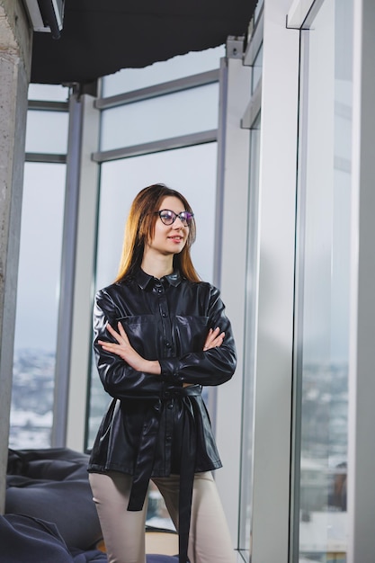 Portrait of a young slender woman in glasses and a black leather shirt Modern woman on the background of the window in the office with a large window