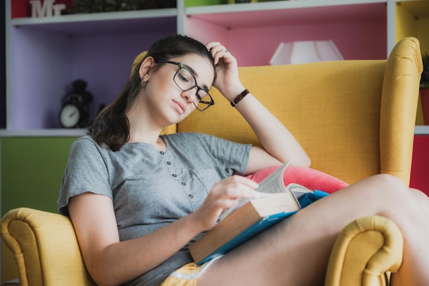 Portrait of young smart and nerdy girl wearing spectacles relaxing on couch at home reading a book while smiling and looking at camera