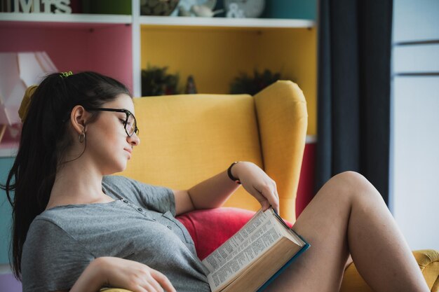 Portrait of young smart and nerdy girl wearing spectacles relaxing on couch at home reading a book while smiling and looking at camera