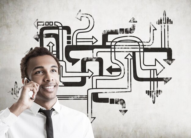 Photo portrait of a young smiling african american businessman with a smartphone standing near a concrete wall with an arrow maze sketch on it.