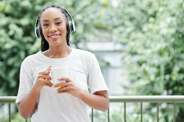 Portrait of young smiling Black woman in headphones standing on balcony with cup of coffee and smiling