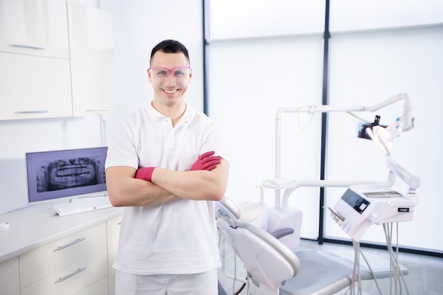 Portrait of a young smiling dentist in work glasses and in a white uniform. The man folded his arms and stands at the dental chair in the office.