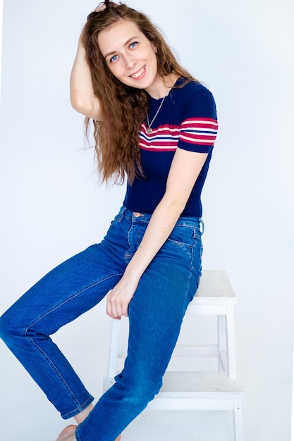 Portrait of a young smiling girl sitting on a chair on a white background with long hair in a tight Tshirt and jeans