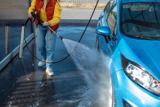 Portrait young, smiling, happy, attractive woman washing automobile at manual car washing self service station, cleaning with foam, pressured water. Transportation, auto, vehicle care concept.