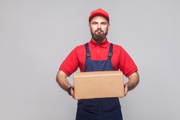 Portrait of young smiling logistic delivery man with beard in blue uniform and red t-shirt standing and holding the cardboard box on grey background. Indoor, studio shot, isolated, copy space.