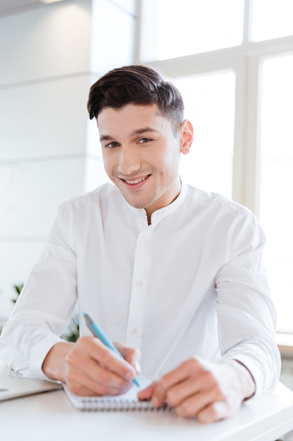 Portrait of young smiling man dressed in white shirt writing notes in notebook. Coworking. Looking at camera.