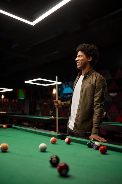 Portrait of young smiling man holding billiards cue stick with pool table on background