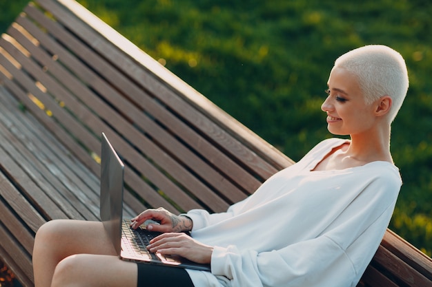 Portrait of young smiling millenial european short haired woman using laptop at bench on green grass...