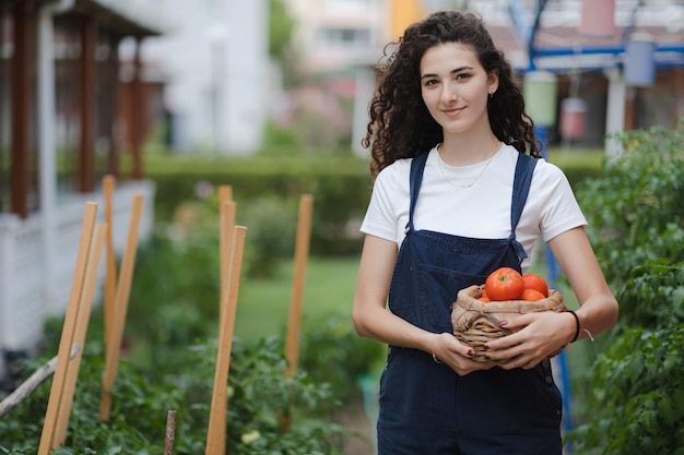 Portrait of young smiling woman with curly hair holding basket with tomatoes looking to camera Countryside living vagetarian healthy eating concept