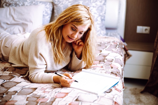 Portrait of young smiling woman with fair hair wearing beige sweater trousers lying on bed writing m