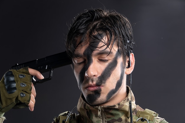 Portrait of young soldier in camouflage stucking gun into his head dark wall