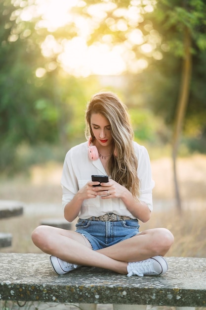 Portrait of a young Spanish woman with hair highlights sitting in a park and texting on her phone