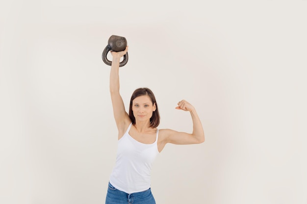 Portrait of young sporty muscular woman with dark hair wearing white top lifting kettle bell showing