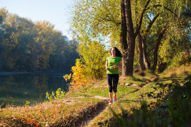 Portrait of young sporty smiling woman jogging early in the morning near the pond. Beautiful landscape of autumn forest and lake with happy woman jogging across