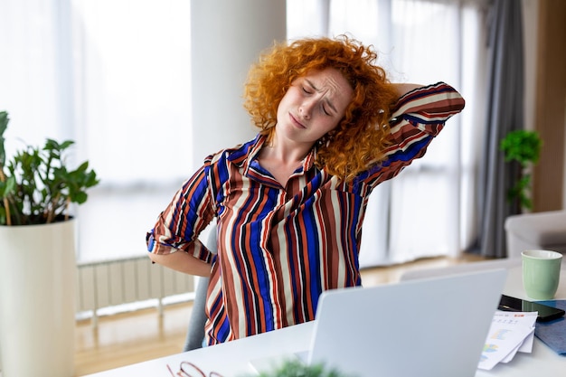 Portrait of young stressed woman sitting at home office desk in front of laptop touching aching back with pained expression suffering from back ache after working on laptop