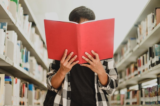 Portrait of a young student in casual style reading a book and covering his face by bookshelf