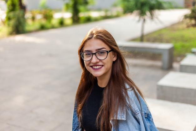 Portrait of a young student woman in a denim jacket and glasses in a park outdoors