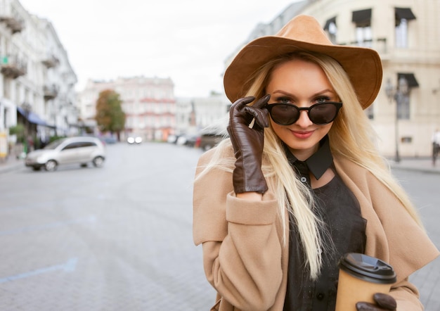 Portrait of young stylish blonde woman in autumn coat, sunglasses, gloves and felt hat holding coffee cup in European city. Beautiful attractive girl