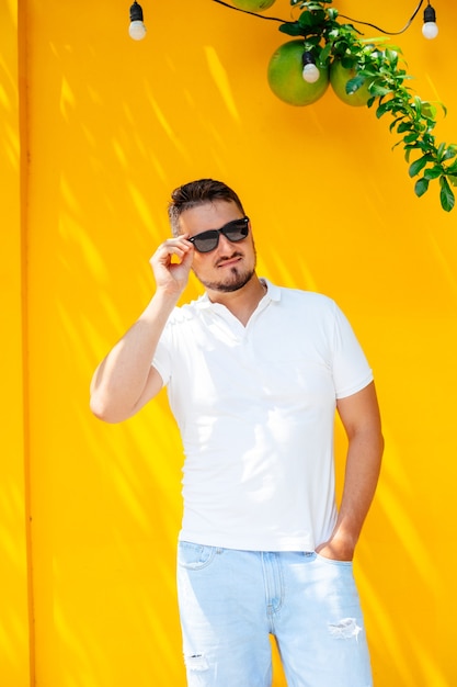 Portrait of a young stylish man with a beard in sunglasses and a white t-shirt on the street against a bright yellow wall, lifestyle. Urban style.