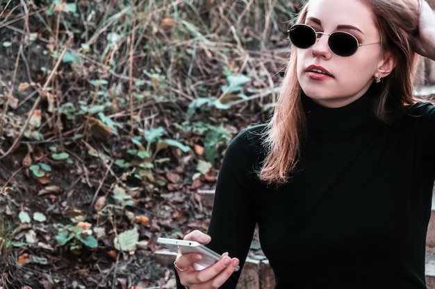 Portrait of young stylish modern woman in sunglasses and black turtleneck sitting on the steps in the park holding mobile phone in her hand. Woman enjoys fresh air, rests from the bustle of the city