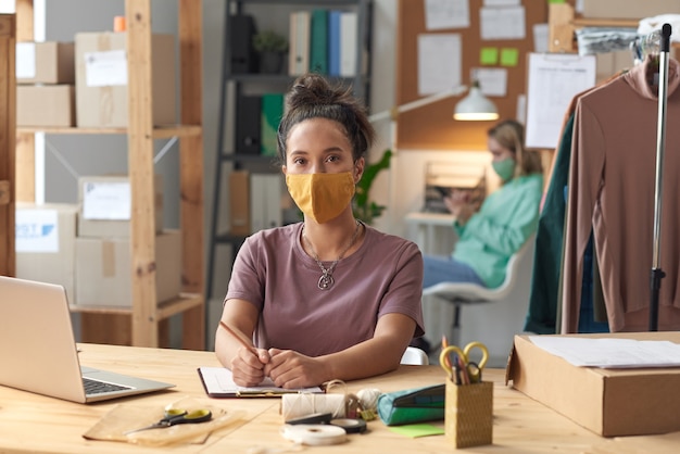 Portrait of young tailor in protective mask looking at front while working at her workplace in workshop