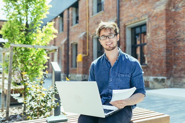 Portrait of a young teacher outside the university campus sitting on a bench with a laptop and smiling