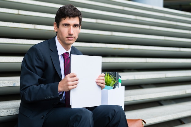 Portrait of young unemployed man looking at camera with sad facial expression while sitting on stairs outdoors after being fired