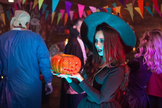 Photo portrait of young witch looking shocked at her punmpkin for halloween celebration. woman with a big witch hat.